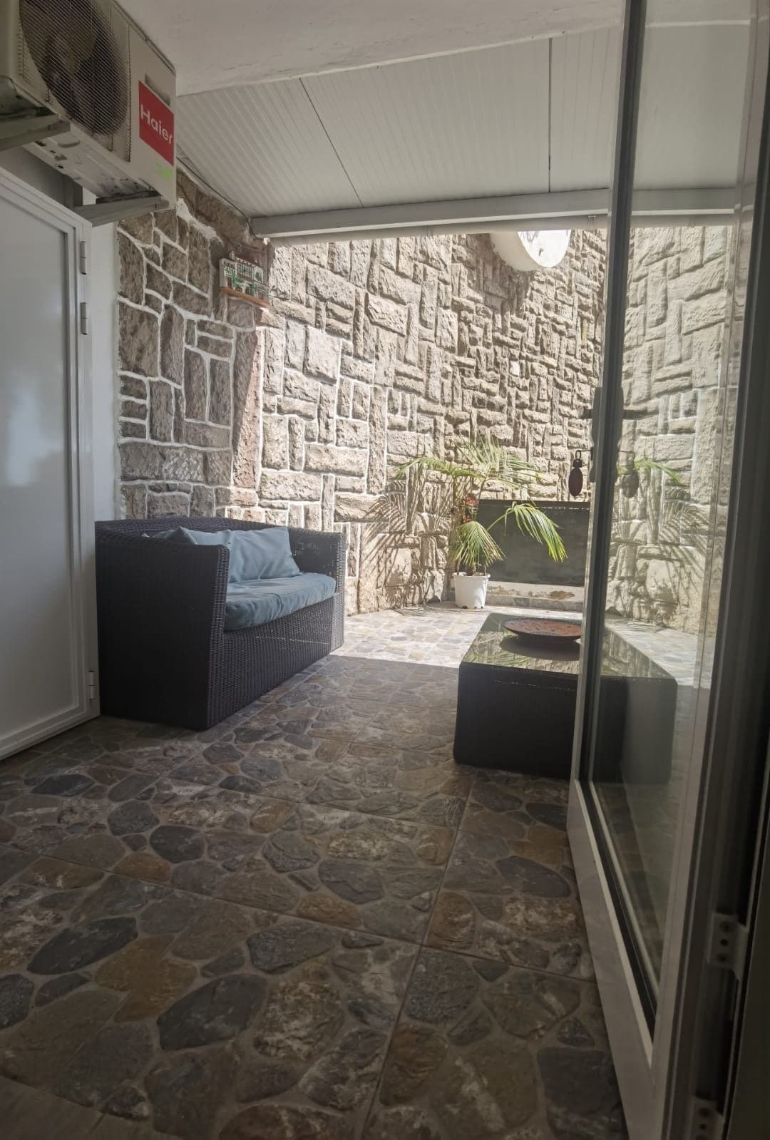 Acogedora terraza cubierta con sofá cómodo, vistas a muros de piedra natural y plantas decorativas. Espacio perfecto para relajarse y disfrutar del ambiente.
